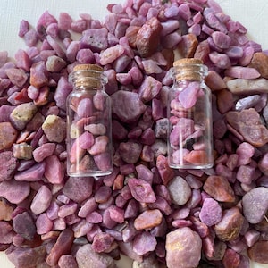 Könnte beinhalten: Zwei kleine Glasflaschen mit Korken, gefüllt mit rosa und lila Edelsteinen, liegen auf einem Bett aus ähnlichen Steinen. Die Edelsteine variieren in Größe und Farbton und erzeugen einen ansprechenden Anblick.