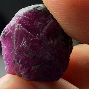 Peut inclure: Une pierre précieuse taillée grossièrement de couleur violet foncé avec une apparence légèrement nuageuse. La pierre précieuse est tenue dans une main.