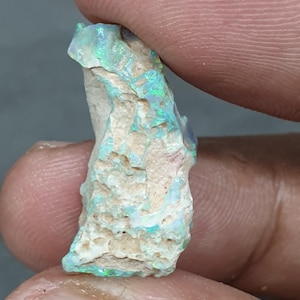 May include: A rough opal gemstone with green and blue flashes, held between fingers. The opal has a textured, off-white base. The gemstone displays iridescent colours.