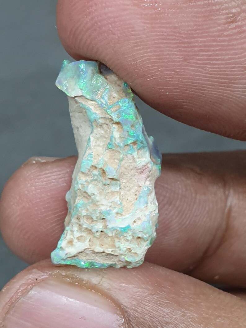 May include: A rough opal gemstone with green and blue flashes, held between fingers. The opal has a textured, off-white base. The gemstone displays iridescent colours.