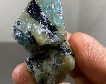 Könnte beinhalten: Ein rohes, unpoliertes Mineralexemplar mit einem durchscheinenden hellblauen Kristallabschnitt, der mit undurchsichtigen schwarzen Turmalineinschlüssen und erdig grünem Matrixgestein mit rauer, natürlicher und facettierter Oberflächenstruktur verwachsen ist.