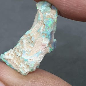 May include: A raw opal gemstone, displaying a mix of blue, green, and white hues. The stone is held between fingers, revealing its natural, irregular form and textured surface. The opal has a rough, unpolished appearance, highlighting its natural beauty.