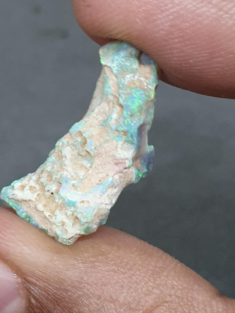 May include: A raw opal gemstone, displaying a mix of blue, green, and white hues. The stone is held between fingers, revealing its natural, irregular form and textured surface. The opal has a rough, unpolished appearance, highlighting its natural beauty.