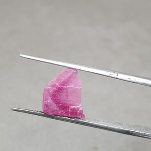 May include: A rough pink gemstone is held by silver tweezers. The gemstone has a textured surface with visible striations and colour variations, ranging from light to deep pink.