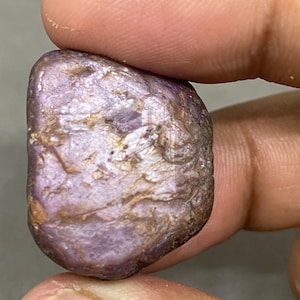 May include: A rough, purple gemstone with a silvery sheen. The gemstone has a natural, irregular shape and is held in a hand.