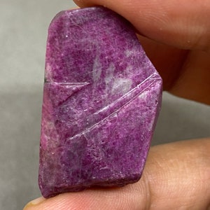 May include: A deep purple, faceted, rough-cut gemstone with white streaks and a textured surface.