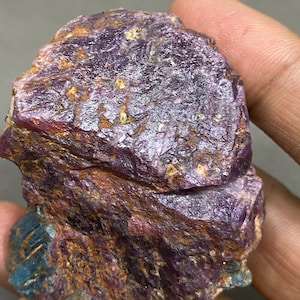 May include: A rough, purple and brown gemstone with a crystalline structure. The gemstone has a natural, unpolished appearance.