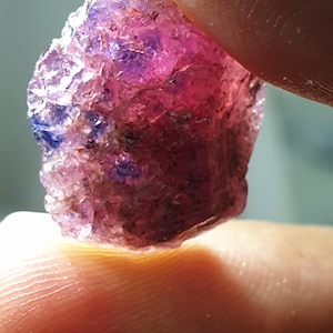 May include: A raw gemstone with a mix of pink, purple, and blue colours. The mineral is held between two fingers, showing its natural, irregular shape and crystalline texture. The gem appears to be a raw sapphire or ruby.