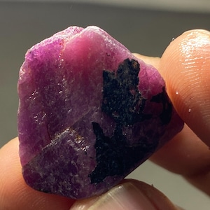 May include: A natural ruby gemstone, roughly cut. The ruby is a deep pinkish-red hue, featuring a dark, black inclusion. The stone has a textured surface and is held between fingers.