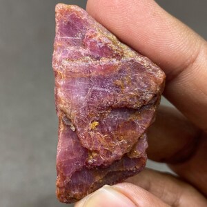 May include: A rough, red ruby gemstone with a layered, crystalline structure. The ruby has a deep red color with hints of orange and gold.