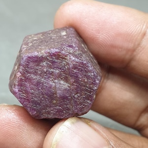Puede incluir: Una piedra preciosa de rubí rojo intenso con forma hexagonal. El rubí es en bruto y sin pulir, con una superficie rugosa.