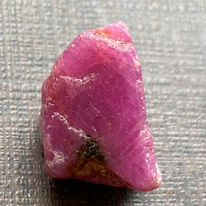 May include: A rough-cut, natural ruby gemstone. The ruby is a deep pinkish-red color with a slightly uneven surface. The gemstone has a raw, unpolished appearance, showcasing its natural crystalline structure. The ruby is a small, handheld size.