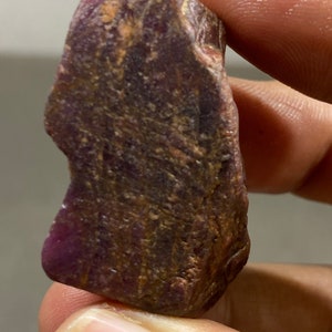 May include: A raw ruby gemstone, displaying a deep burgundy hue with a textured surface. The stone has areas of lighter brown and tan, and is held between two fingers.