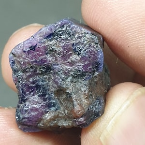 May include: A rough-cut, natural gemstone with a mix of purple, blue, and gray hues. The stone has a textured surface and irregular shape, showcasing its raw, unpolished state. The gemstone is held between fingers, highlighting its size and detail.
