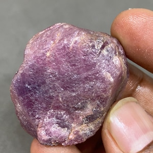 May include: A rough, uncut, purplish-pink gemstone. The natural stone has a textured surface with variations in color and tone. The gemstone is held between two fingers, showcasing its size and shape. The stone appears to be a raw mineral specimen.