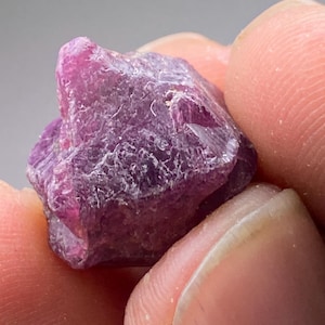 Könnte beinhalten: Ein rauer, ungeschliffener Rubin-Edelstein, der zwischen Fingern gehalten wird. Der Rubin hat eine tiefe, lebendige, rötlich-rosa Farbe mit einer strukturierten, unebenen Oberfläche. Der Edelstein ist ein natürliches, rohes Mineral.