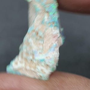 May include: A raw opal gemstone, displaying a creamy white base with iridescent flashes of blue, green, and pink. The stone's fractured surface reveals its natural texture. The opal is held between two fingers.