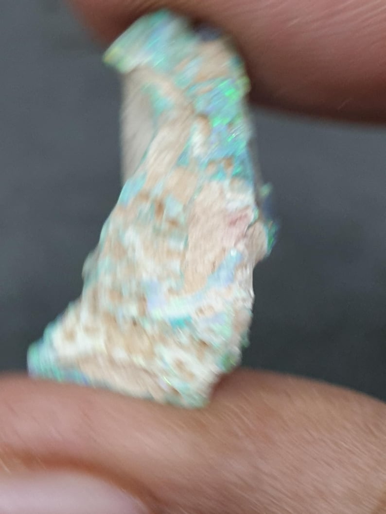 May include: A raw opal gemstone, displaying a creamy white base with iridescent flashes of blue, green, and pink. The stone's fractured surface reveals its natural texture. The opal is held between two fingers.