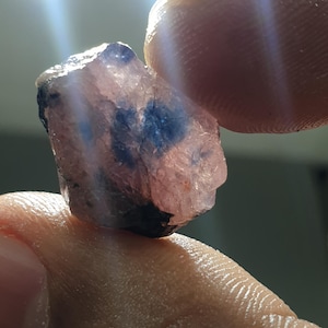 May include: A raw gemstone, exhibiting a pinkish-purple colour with dark blue patches. The stone is held between fingers, with light highlighting its textured surface. It appears to be a natural, unpolished mineral sample.