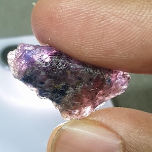 May include: A raw gemstone, displaying a mix of purple and pink colours, held between two fingers. The stone has a textured surface and appears translucent. The shape is irregular, highlighting its natural form and characteristics.