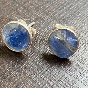 May include: A pair of silver stud earrings with blue gemstone accents. The gemstones are round and have a cloudy, translucent appearance.