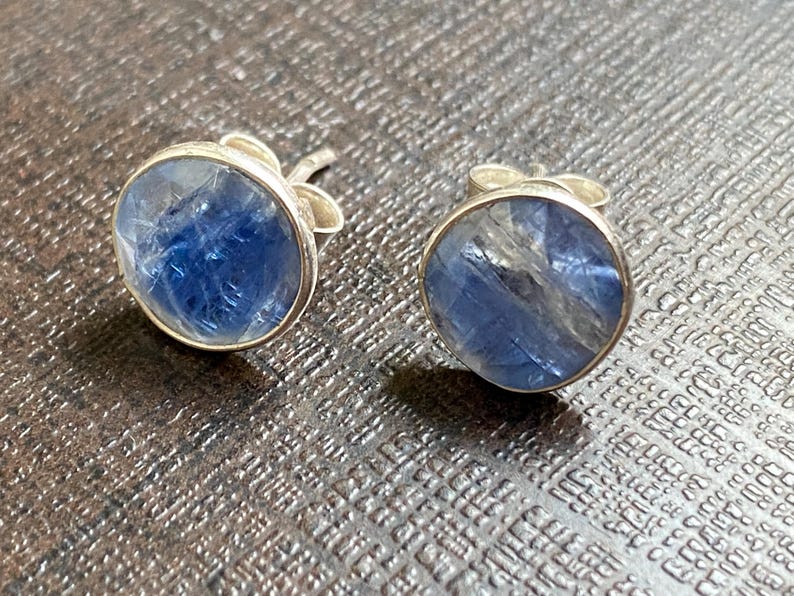May include: A pair of silver stud earrings with blue gemstone accents. The gemstones are round and have a cloudy, translucent appearance.