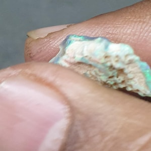 May include: A close-up of a rough opal gemstone. The opal shows a mix of colours, including blue, green, and hints of pink, with a textured, porous surface. The gemstone is held between fingers.