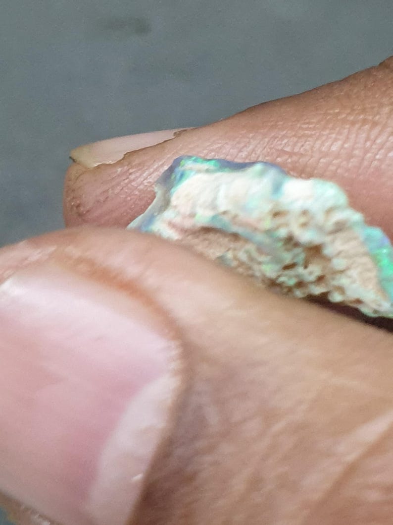 May include: A close-up of a rough opal gemstone. The opal shows a mix of colours, including blue, green, and hints of pink, with a textured, porous surface. The gemstone is held between fingers.