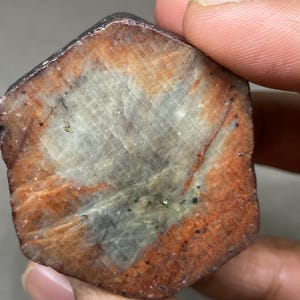 May include: A rough, irregularly shaped, dark grey and brown gemstone with a lighter grey, almost white, centre. The gemstone has a striated pattern and appears to be translucent.