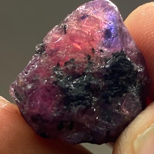 May include: A rough, purple and pink gemstone with black inclusions. The gemstone is irregularly shaped and has a natural, unpolished finish.