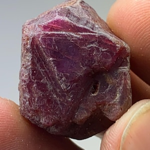 Puede incluir: Un cristal de rubí rojo intenso con una superficie rugosa y facetada. El cristal es una gema natural con un color rico y vibrante.