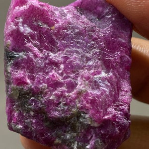 May include: A rough, pink and purple gemstone with a dark grey, almost black, inclusion. The gemstone is irregularly shaped and has a rough, natural texture.