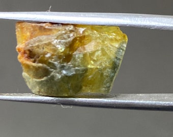 May include: A rough, green and yellow gemstone with a faceted surface, held in tweezers.