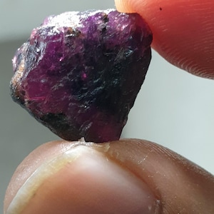 May include: A raw, unpolished gemstone in a deep purple colour, held between two fingers. The stone has a crystalline structure with dark inclusions, reflecting light. The overall shape is irregular and natural.