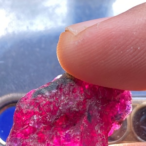 May include: A large, rough, pink ruby gemstone with a dark grey inclusion. The ruby is being held in a hand.