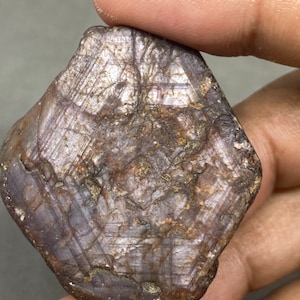 May include: A rough, dark purple gemstone with a hexagonal shape and a striated surface. The gemstone has a natural, unpolished finish.