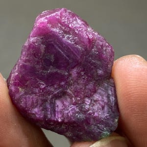 May include: A large, rough, pink ruby gemstone with a deep red hue. The ruby has a natural, unpolished finish and is a beautiful example of this precious stone.