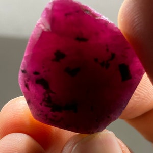 Può includere: Una pietra preziosa rosa intenso con inclusiones nere. La pietra preziosa è sfaccettata e ha una forma irregolare e ruvida.