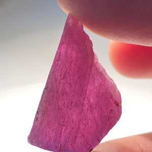 May include: A raw, pink gemstone with a rough, uneven surface. The stone is held between two fingers, highlighting its natural, unpolished texture and vivid colour. The gem is a raw mineral specimen, with a translucent quality.