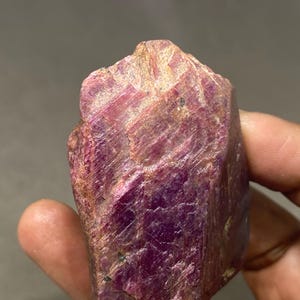 May include: A rough-cut ruby gemstone with a deep red-purple hue. The natural stone has a textured surface with visible lines and variations in color, ranging from light pink to dark crimson. The ruby is held in a hand, showcasing its raw, unpolished form.