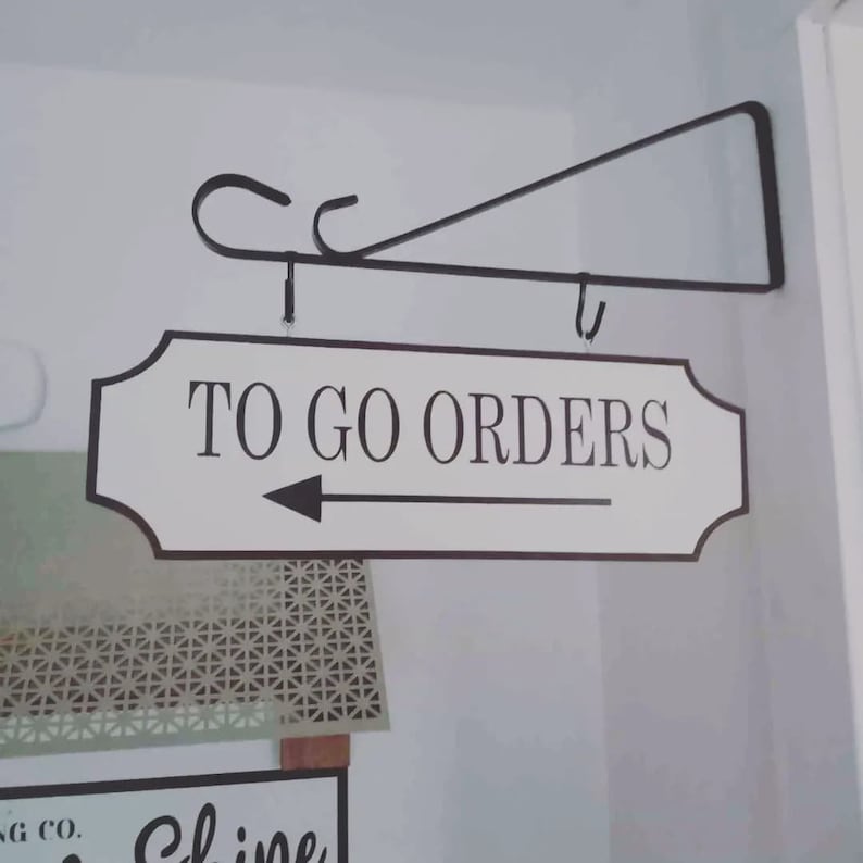 May include: White sign with black lettering and an arrow pointing to the left. The sign reads "To Go Orders". The sign is hanging from a black metal bracket.
