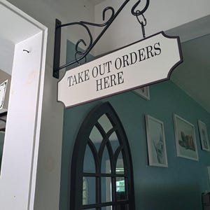 May include: A black metal hanging sign with the text "TAKE OUT ORDERS HERE" in black letters on a white background. The sign is suspended from a decorative bracket attached to a wall.