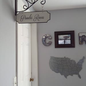 May include: A white sign with black lettering that reads "Powder Room" is hanging from a black metal hook on a white wall. The sign is attached to the wall with a black metal bracket. The sign is in a hallway with a white door and a light switch on the wall.
