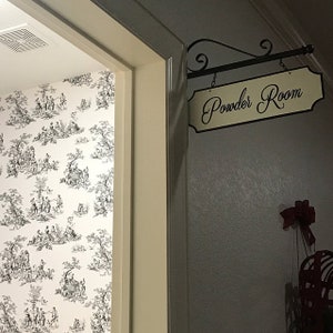 May include: A white door with a black and white patterned wallpaper behind it. A sign above the door reads "Powder Room" in black cursive lettering on a cream background.