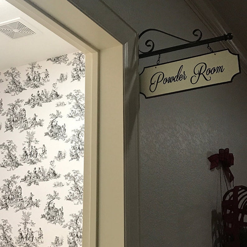 May include: A white door with a black and white patterned wallpaper behind it. A sign above the door reads "Powder Room" in black cursive lettering on a cream background.