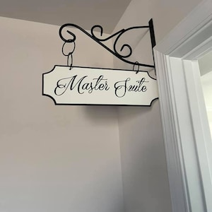 May include: A white sign with black lettering that reads "Master Suite" hanging from a black metal sign holder.