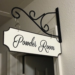 May include: A white sign with black lettering that reads "Powder Room" hanging from a black metal hook. The sign is attached to a white wall.