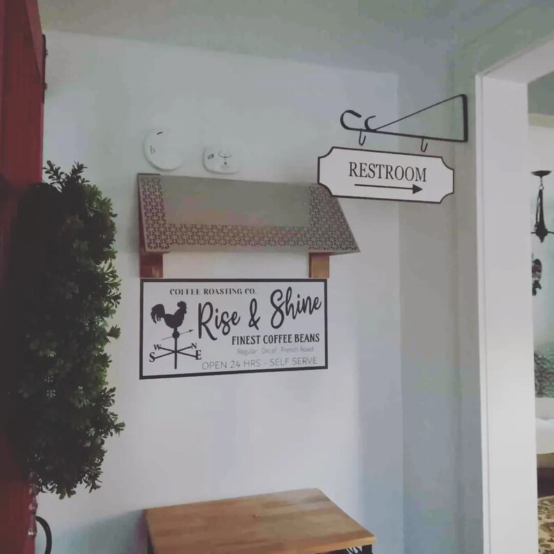 May include: A white sign with black text that reads "COFFEE ROASTING CO. Rise & Shine FINEST COFFEE BEANS Regular Decaf French Roast OPEN 24 HRS - SELF SERVE" with a black rooster illustration and a compass rose. A white sign with black text that reads "RESTROOM" with a black arrow pointing to the right.