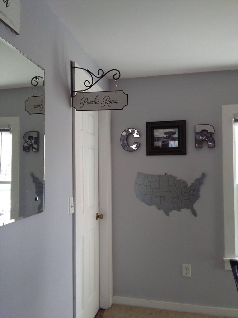 May include: A white door with a black sign that says "Powder Room" hanging above it. The sign is attached to a black metal bracket. There is a gray map of the United States on the wall to the right of the door. There are also three silver letters, "C", "O", and "R", on the wall.