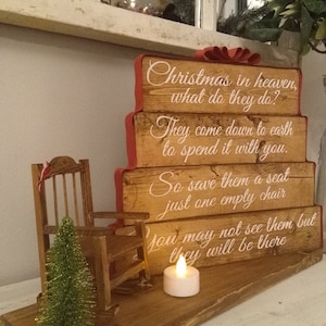May include: Wooden Christmas sign with a red bow and the text "Christmas in heaven, what do they do? They come down to earth to spend it with you. So save them a seat just one empty chair You may not see them, but they will be there". A rocking chair and a small pine tree are in front of the sign.